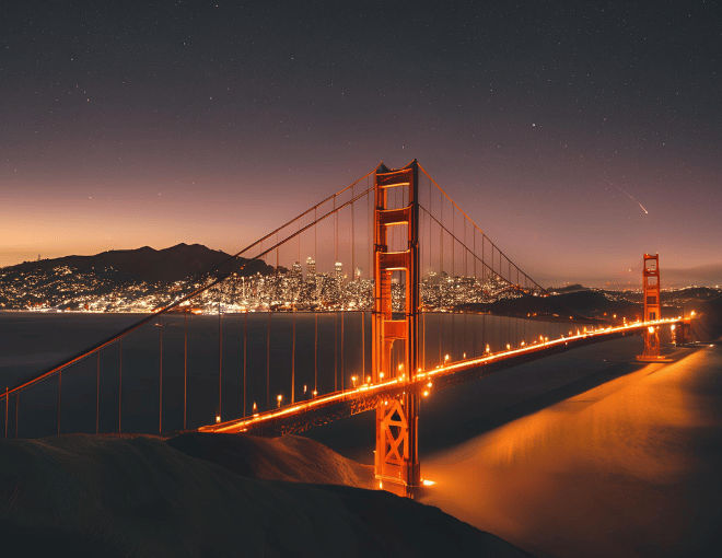Golden Gate Bridge glows under the night sky, city lights twinkling in the distance, reflecting on the bay's calm waters, creating a mesmerizing San Francisco skyline.
